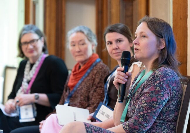 Liz Collins, Executive Director of Pharmaceutical Sciences at Pfizer, speaks into a microphone at the Developing Business-Aware Academics launch event. Behind her are sat Flora Hamilton, Paula Whitehouse, and Danielle Croucher