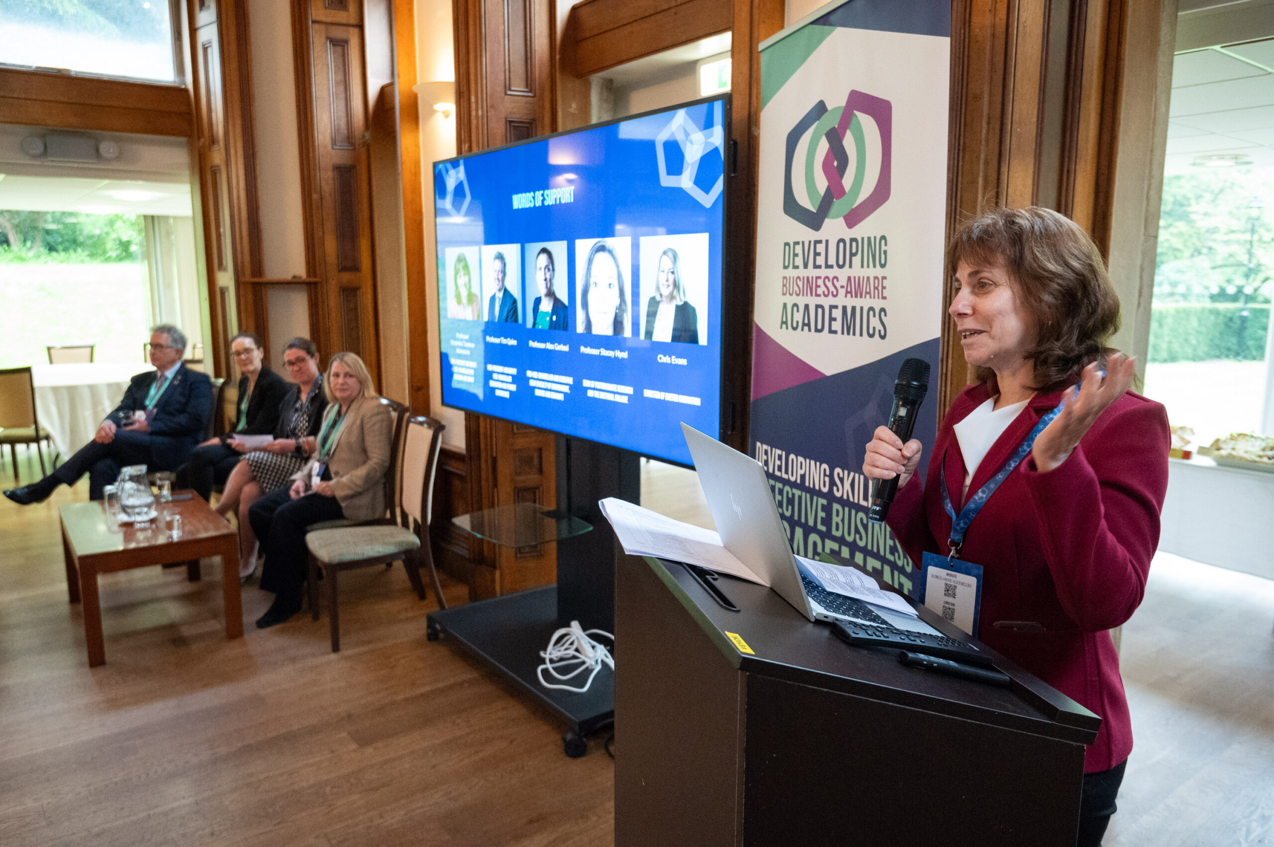 Professor Krasimira Tsaneva-Atanasova​ presents from a podium in front of a Developing Business-Aware Academics banner. 4 people watch from the other side of the screen