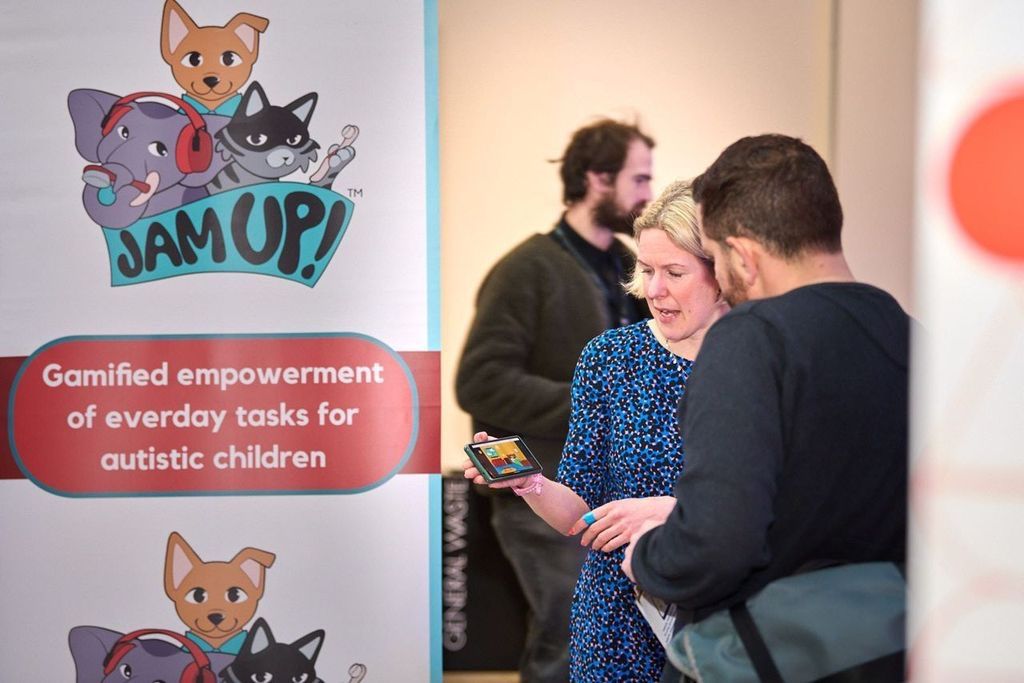 Rachael Malthouse stands in front of a banner for Jam Up!, showing the app on her phone to a man. The banner features a logo of three cartoon animals, and the text 