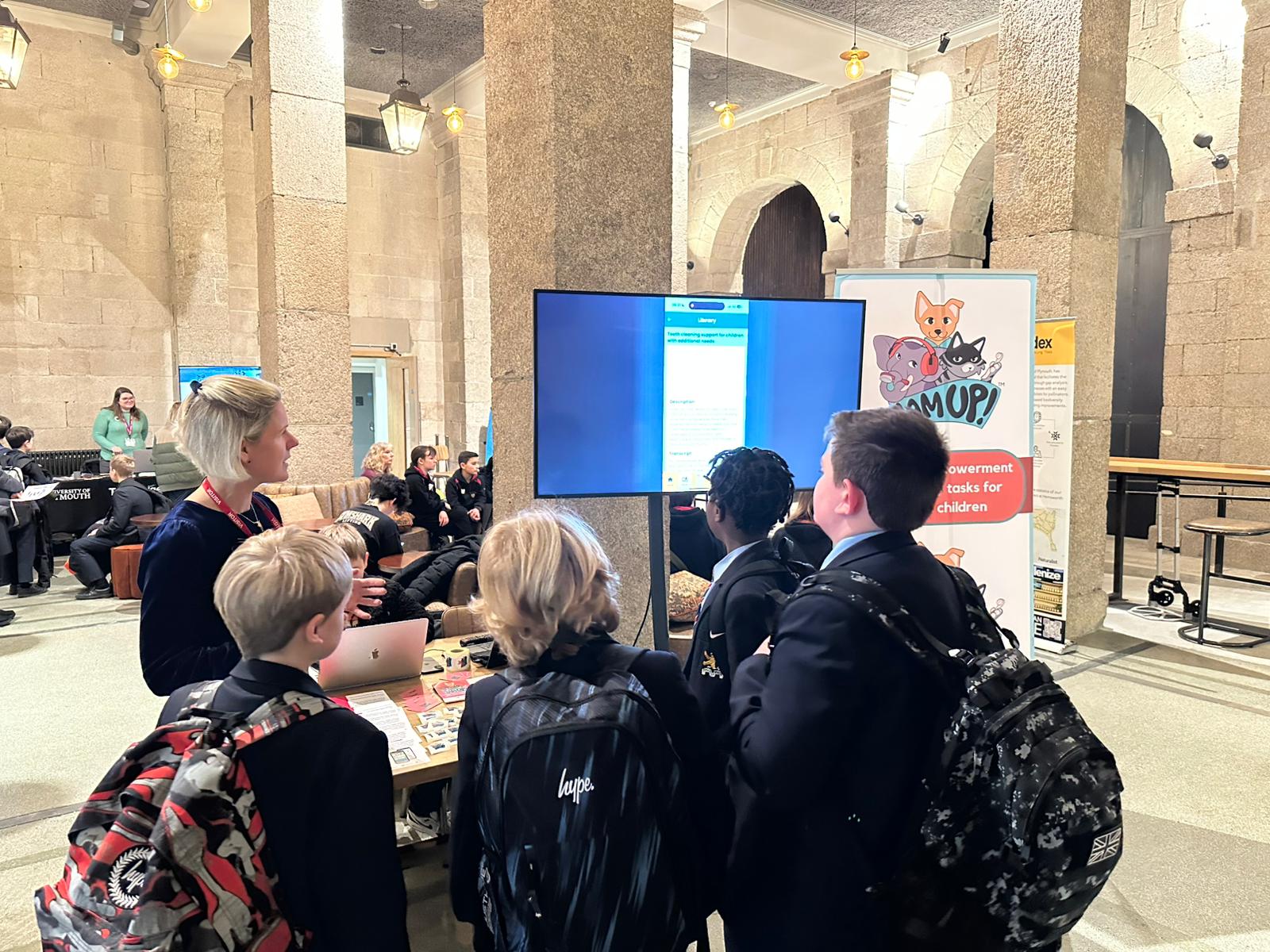 A photo of Rachael Malthouse in front of a stand for Jam Up! She is talking to a group of young boys about the app
