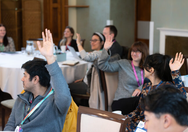 A photo of three groups of people in round table discussions. A number of the people have raised their hands